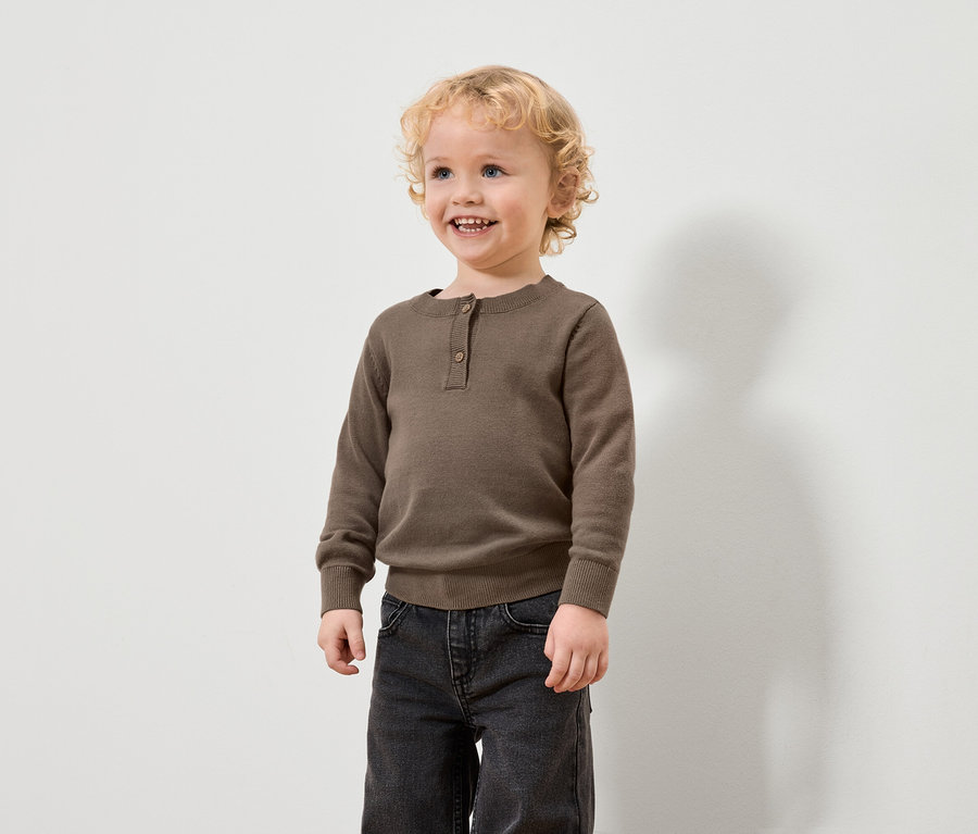 Ein Junge mit blonden Locken trägt einen braunen Pullover und schwarze Jeans.