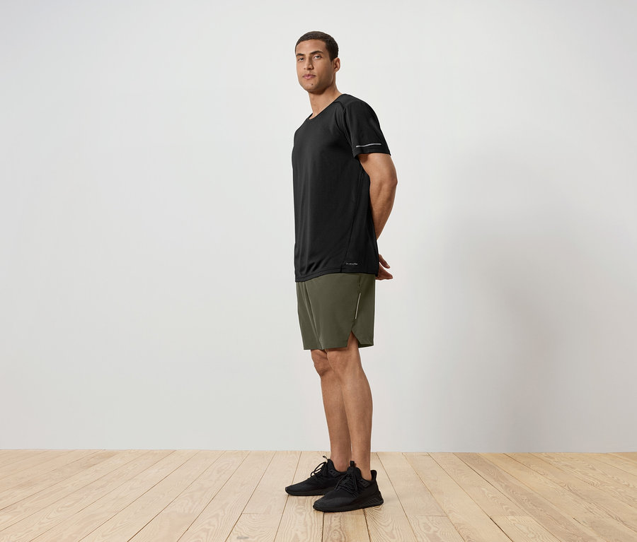 Seitenansicht eines Mannes in einem schwarzen T-Shirt, grünen Shorts und schwarzen Turnschuhen, der auf einem Holzboden vor einer weißen Wand steht.