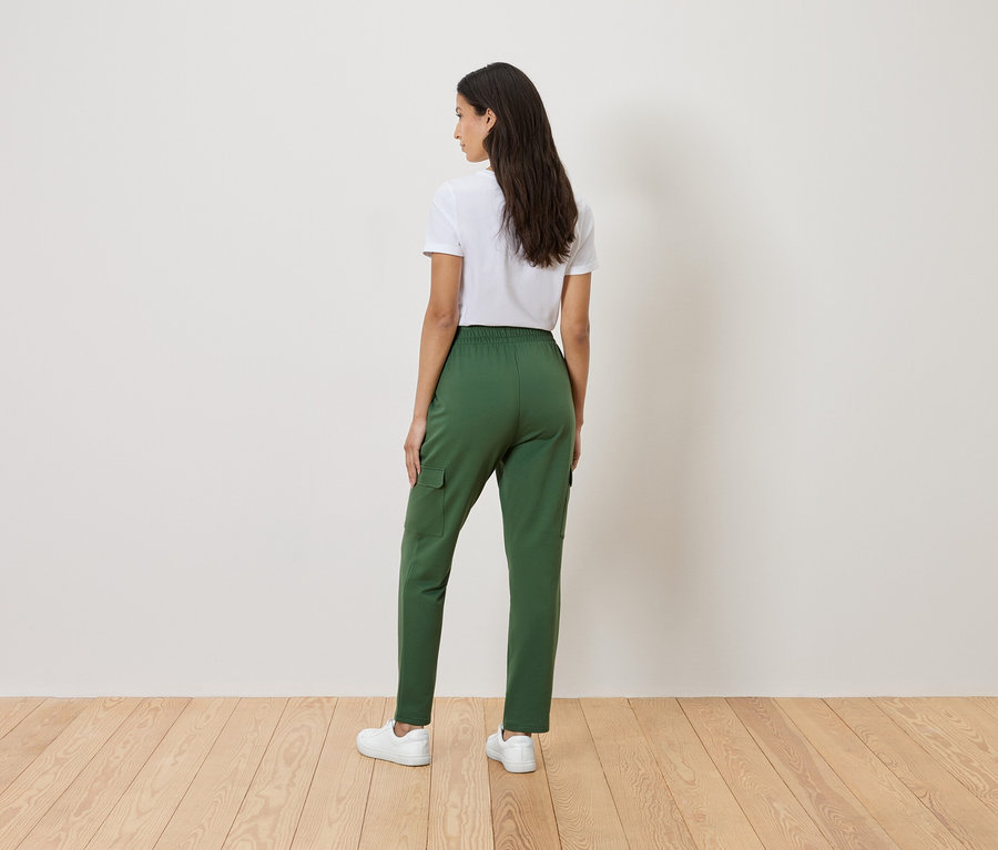 Eine Frau steht mit dem Rücken zur Kamera und trägt ein weißes T-Shirt, eine grüne Hose und weiße Turnschuhe.