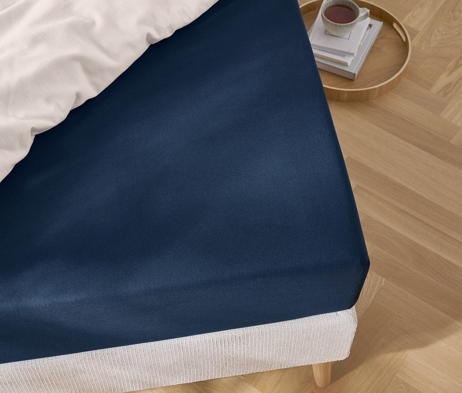 Detailansicht eines blauen Spannbettlakens auf einem Bett mit weisser Bettwäsche und einem Tablett mit Büchern und einer Tasse.