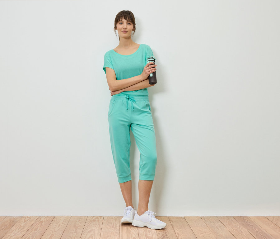 Frau in mintfarbenem Shirt und Caprihose steht mit verschränkten Armen und hält eine Wasserflasche.