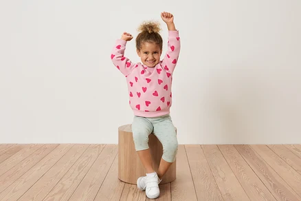 Ein junges Mädchen sitzt auf einem Holzhocker, die Arme hochgestreckt, und trägt ein rosa Sweatshirt mit roten Herzen und hellgrüne Leggings.