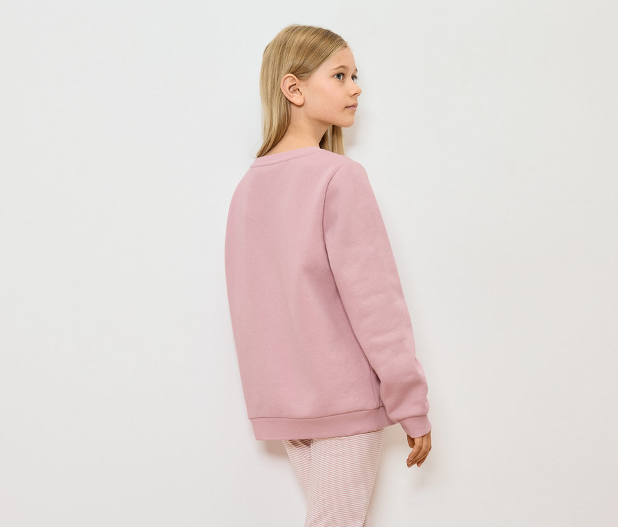 Ein junges Mädchen steht seitlich in einem rosa Pullover und gestreiften Hosen vor einer weißen Wand.