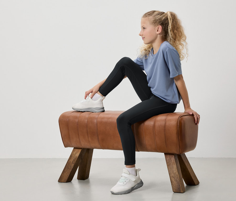 Ein junges Mädchen mit blonden, zu einem Pferdeschwanz gebundenen Haaren sitzt auf einem Turnbock. Sie trägt ein blaues T-Shirt, schwarze Leggings und weiße Turnschuhe.