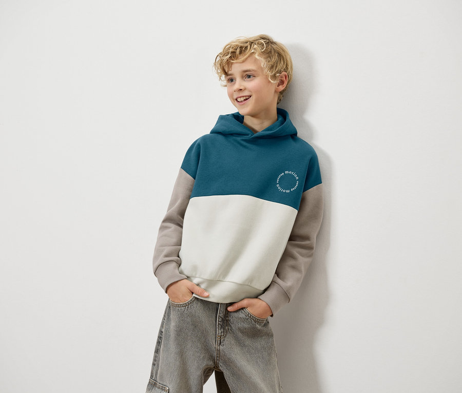 Ein blonder Junge lehnt an einer Wand und trägt einen blau-weiß-braunen Kapuzenpullover und graue Jeans.