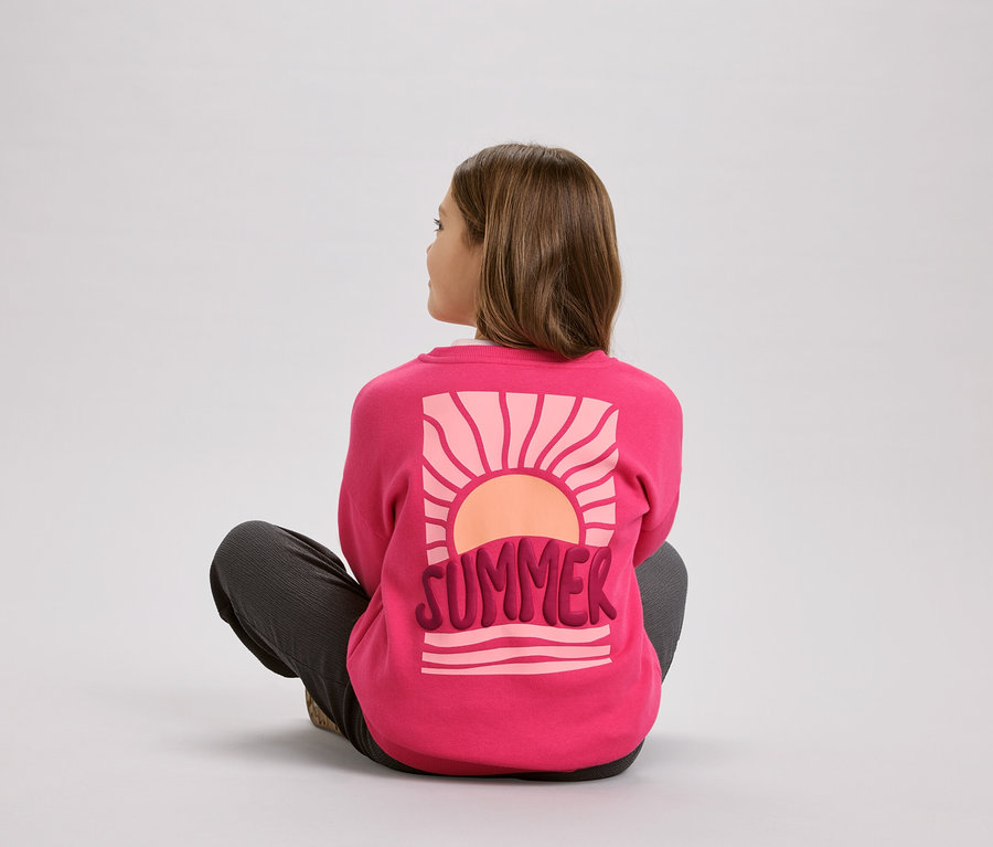 Mädchen sitzt im Schneidersitz und trägt einen pinken Pullover mit dem Aufdruck "Summer" und einem Sonnenmotiv.