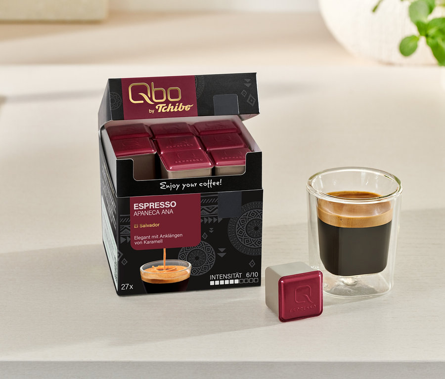 Eine geöffnete Packung Qbo Espresso APANECA ANA mit 27 Kapseln neben einem Glas Espresso und einer einzelnen Kapsel.