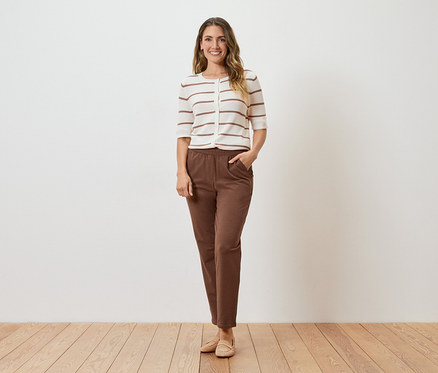 Eine Frau steht vor einer weißen Wand und trägt eine gestreifte Strickjacke, braune Hosen und beigefarbene Schuhe.