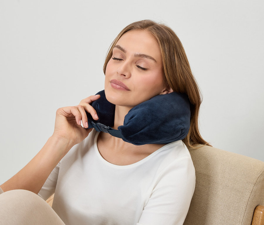 Frau mit geschlossenen Augen trägt eine dunkelblaue Nackenstütze, während sie in einem Sessel sitzt.