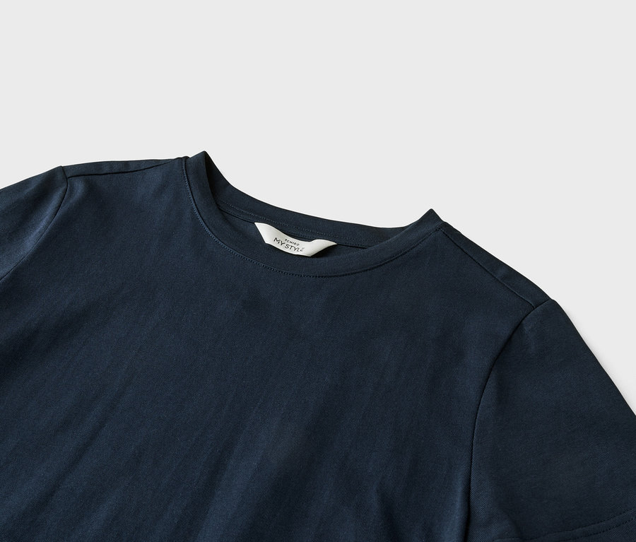 Nahaufnahme eines marineblauen T-Shirts mit einem weißen Etikett mit der Aufschrift "ICHIBO MY STYLE" am Kragen, das auf einem hellgrauen Hintergrund liegt.