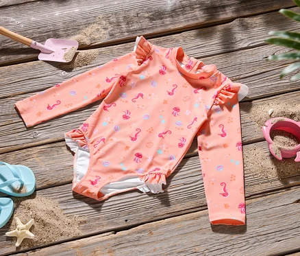 Ein rosa Badeanzug mit Meeresmotiven liegt auf einem Holztisch, zusammen mit einer blauen Flip-Flop, einer rosa Schaufel und einem rosa Sandförmchen.