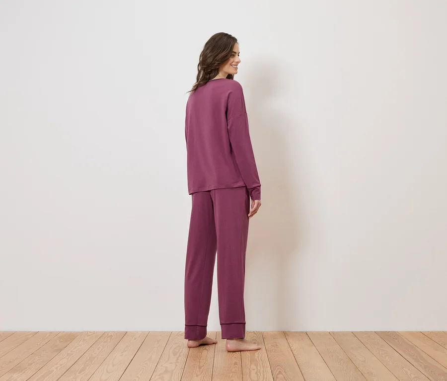 Frau von hinten in langärmligem, weinrotem Pyjama-Set auf Holzboden vor einer weißen Wand.