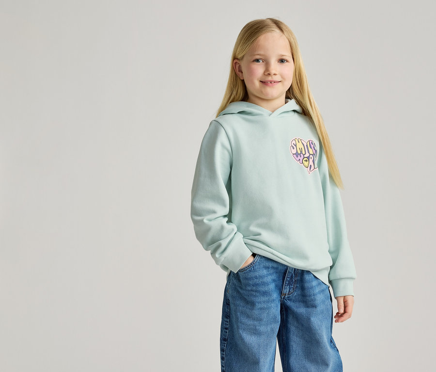 Mädchen mit blonden Haaren, trägt einen hellblauen Hoodie mit Herz-Aufdruck und Jeans.