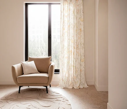 Ein beiger Sessel mit einem weißen Kissen steht vor einem großen Fenster, daneben hängt ein SCHÖNER WOHNEN-Kollektion Vorhang mit verdeckten Schlaufen »LOVELY« mit Blumenmuster auf einem runden Teppich.
