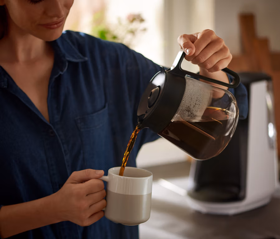 Frau gießt Kaffee aus einer Kanne in eine Tasse. Im Hintergrund ist eine »Let's Brew« Filterkaffeemaschine zu sehen.