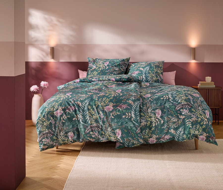 Ein Bett mit grünem Blumenmuster-Bettbezug und zwei Kissen in einem Schlafzimmer mit horizontal gestreiften rosa-burgunderroten Wänden. Zwei Wandleuchten beleuchten die Wand über dem Bett. Eine weiße Vase mit rosa Blumen steht links, ein Holznachttisch mit Büchern und einer Kerze rechts. Ein heller Teppich liegt auf dem Holzboden.