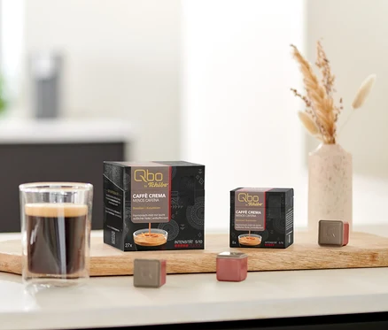 Auf einem Tisch stehen Qbo Caffè MENOS CAFEÍNA Kapselpackungen und eine Tasse Kaffee auf einem Holzbrett.