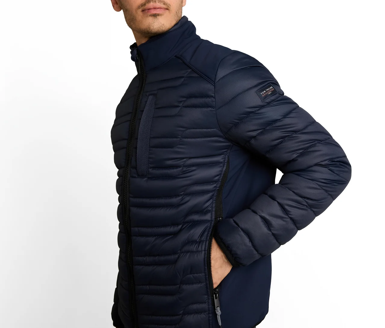Mann trägt eine dunkelblaue Steppjacke mit Stehkragen, seine rechte Hand steckt in einer Seitentasche. Ein "Tom Tailor"-Patch ist am linken Ärmel.
