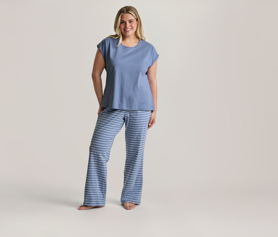 Lächelnde blonde Frau steht barfuß in einem Pyjama-Set mit blauem T-Shirt und blau-weiß gestreiften Hosen.