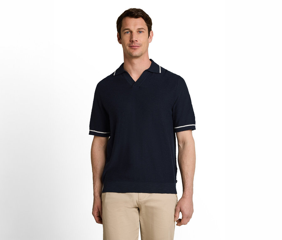 Ein Mann steht frontal da und trägt ein dunkelblaues, kurzärmeliges Poloshirt mit weißen Streifen am Kragen und an den Ärmeln sowie eine hellbraune Hose.