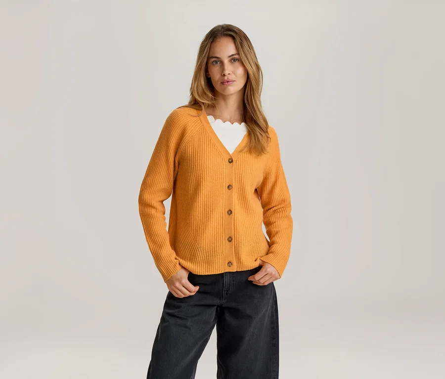 Frau mit langen hellbraunen Haaren, die mit den Händen in den Taschen steht und einen offenen orangefarbenen Merino-Cardigan über einem weißen Oberteil mit Wellenkante und einer dunklen weiten Hose trägt.