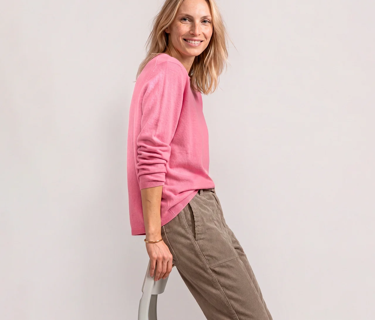 Eine lächelnde Frau trägt einen roséfarbenen Street One Strickpullover und beigefarbene Cordhosen, während sie sich auf einen Stuhl lehnt.