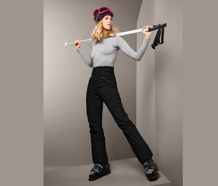Blonde Frau in grauem Shirt und schwarzer High-Waist-Softshell-Skihose hält Skistöcke.