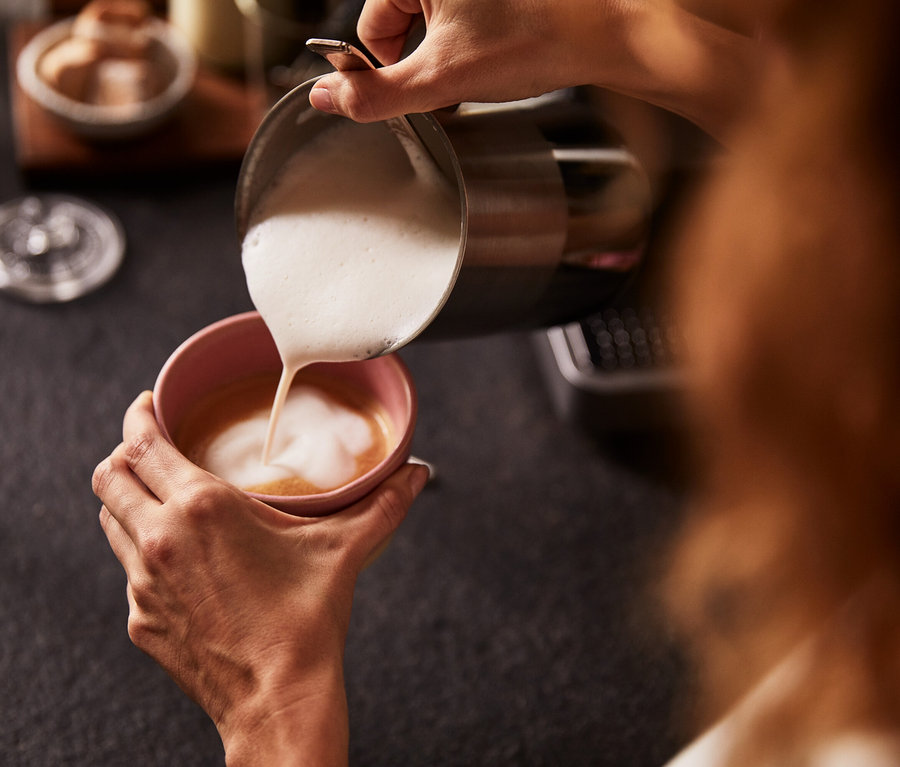 Frau gießt geschäumte Milch aus einem Hand-Milchaufschäumer in eine rosa Tasse Kaffee.