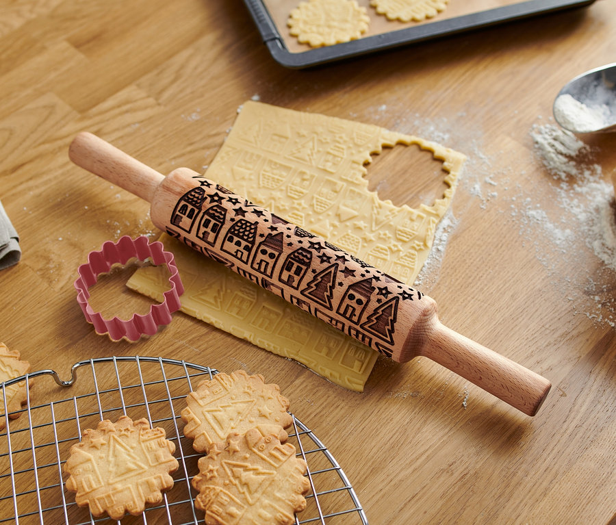 Auf einem Holztisch liegen eine Motiv-Teigrolle, ein Ausstecher und gebackene Kekse.