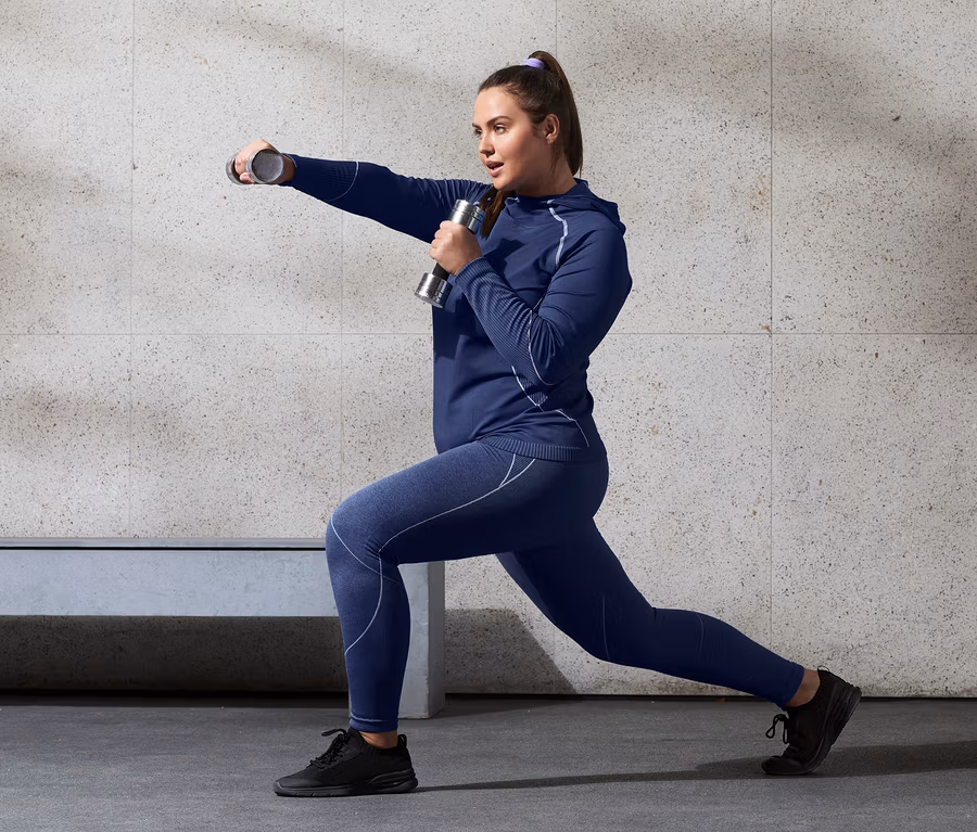 Frau macht Ausfallschritte mit Hanteln, sie trägt ein Seamless-Funktionsshirt mit Kapuze und Seamless-Sporttight.