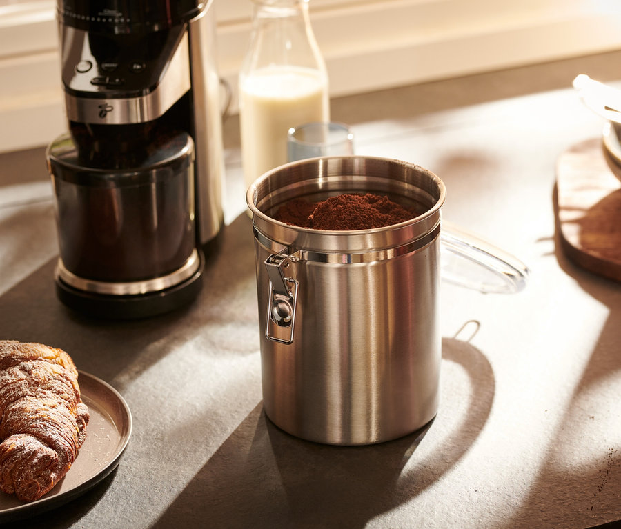 Eine Aromadose aus Edelstahl steht auf einer Küchentheke neben einer Kaffeemaschine und einem Teller mit einem Croissant.