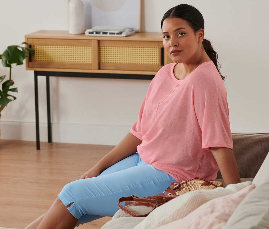 Eine Frau sitzt auf einem Sofa und trägt einen Feinstrick-Pullover aus Leinenmix, eine hellblaue Bengalin-Hose in 3/4-Länge und Espadrilles. Auf dem Sofa liegt eine Beuteltasche.