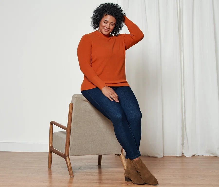 Frau sitzt auf einem Stuhl und trägt einen Strickpullover, Jeggings und Stiefelette aus Veloursleder.