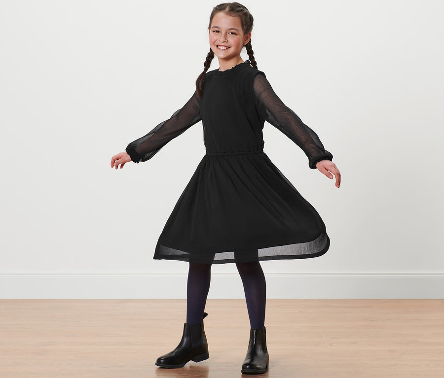 Lächelndes Mädchen mit Zöpfen steht in schwarzem Chiffonkleid.