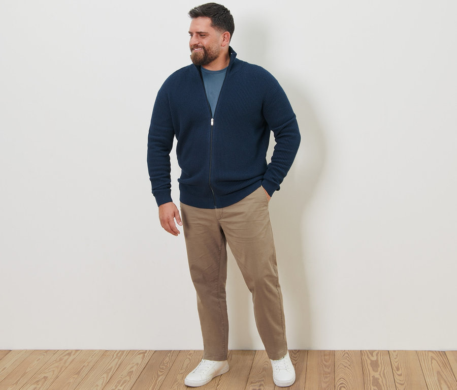 Mann steht in sandfarbenen Chinos, 2 Langarmshirts und marineblauer Strickjacke.