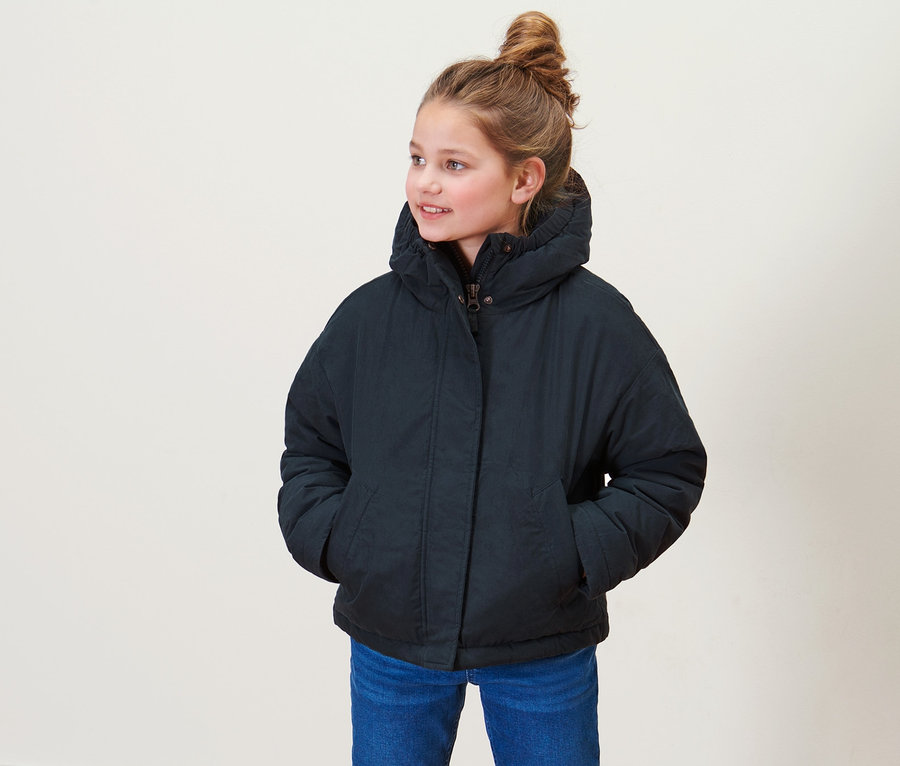 Mädchen steht und trägt eine Kinder-Übergangsjacke, blaue Jeans und braune Schuhe.