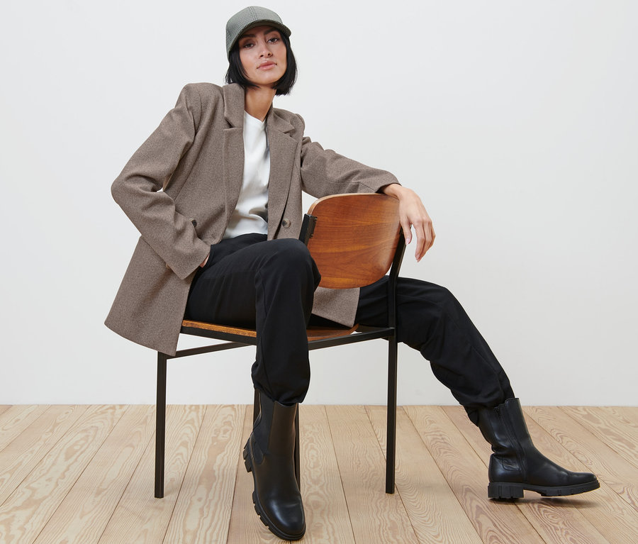 Frau sitzt auf einem Stuhl und trägt einen Blazer, Hose und Stiefel.