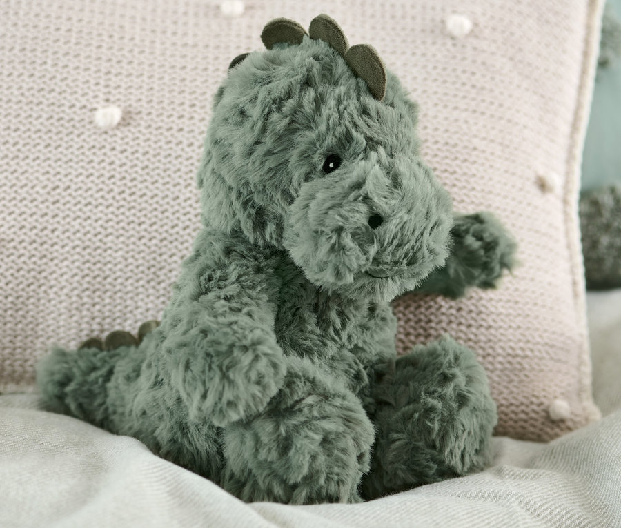 Ein Plüschtier Dino sitzt auf einem Bett neben einem beige Kissen.