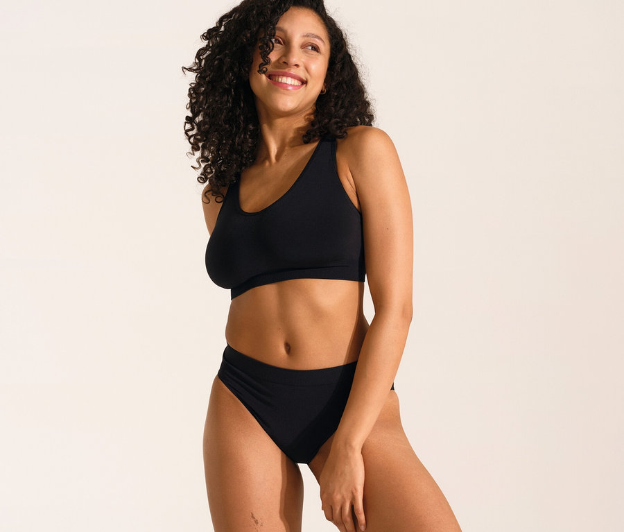 Lachende Frau mit lockigem Haar trägt schwarzen SCHIESSER Soft BH mit Pads und schwarzen SCHIESSER Rio Slip.