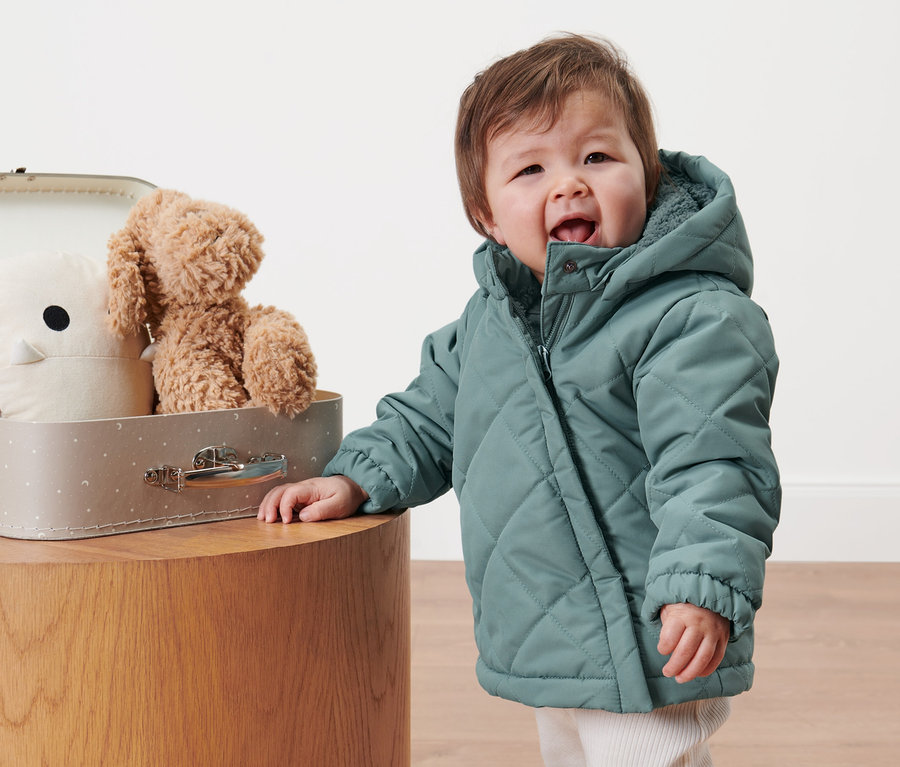 Kleinkind in Kleinkind-Steppjacke steht neben einem Koffer mit Spielzeug.