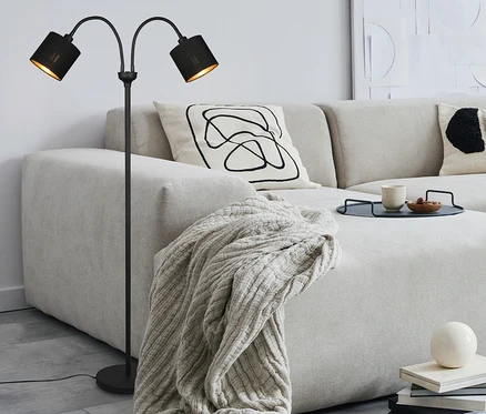 Eine schwarze Reality Stehleuchte »Tommy« steht neben einem beigen Sofa mit Kissen und einer Decke.