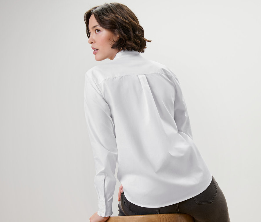 Frau sitzt seitlich auf einem Holzstuhl, trägt eine weiße Bluse mit Rüschendetail.