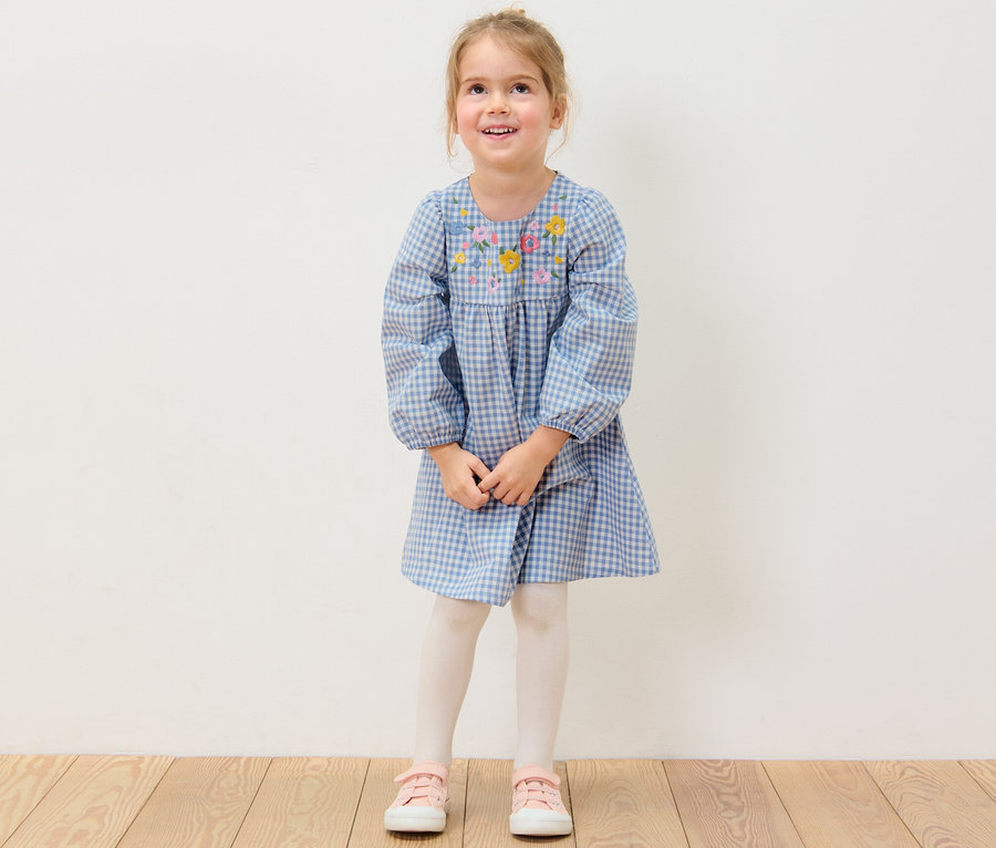 Ein lächelndes Mädchen trägt ein gewebtes Kinderkleid mit Blumenstickerei.