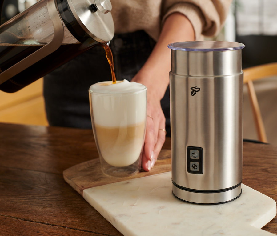 Frau gießt Kaffee in ein Glas mit Milch neben einem elektrischen Milchaufschäumer aus Edelstahl.