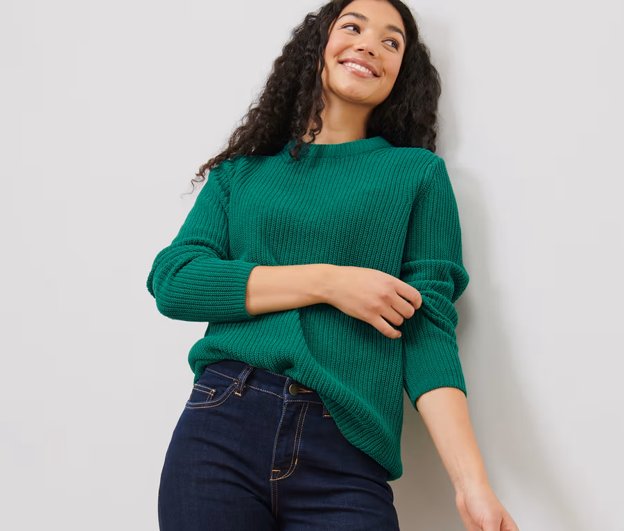 Eine Frau mit dunklen lockigen Haaren trägt einen grünen Grobstrickpullover und Jeans.