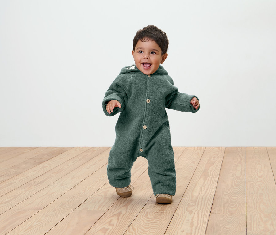 Lächelndes Kleinkind in grünem Overall mit Kapuze und Schuhen auf Holzboden.