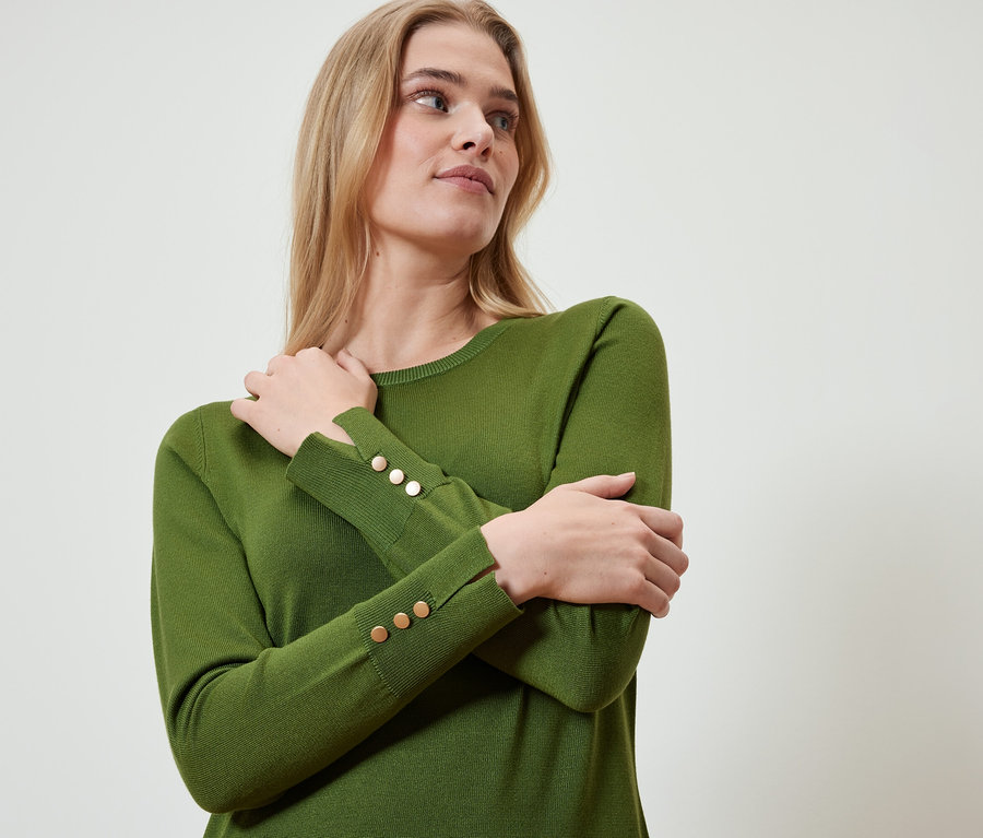 Blonde Frau trägt einen olivgrünen Pullover mit goldenen Knöpfen an den Ärmeln, die Arme sind verschränkt.