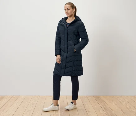 Eine Frau posiert in einem langen, marineblauen Steppmantel, marineblauen Hosen und weißen Turnschuhen.