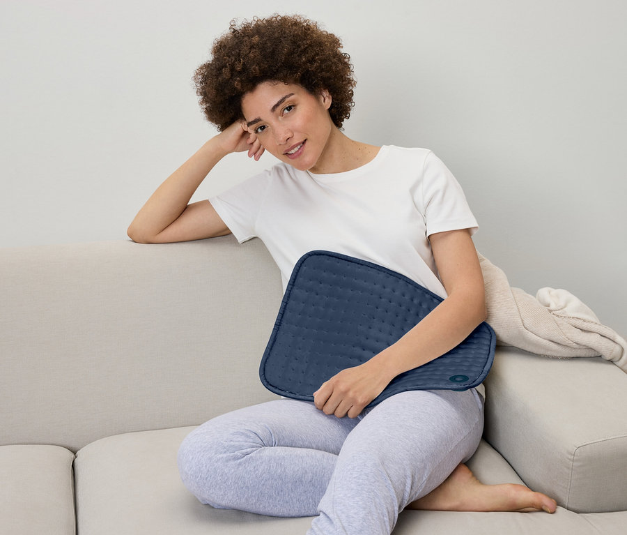 Frau sitzt auf der Couch und hält ein blaues Heizkissen auf dem Schoß.