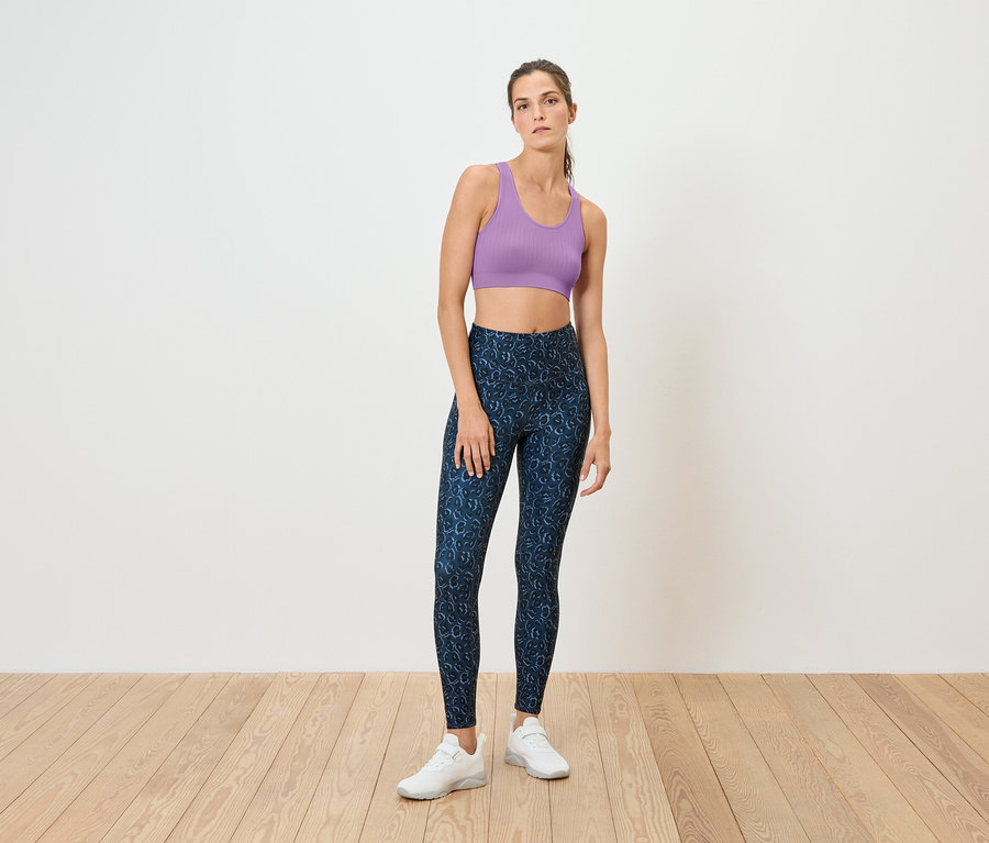 Eine Frau steht auf einem Holzboden und trägt einen lilafarbenen Sport-BH und blaue Leggings.